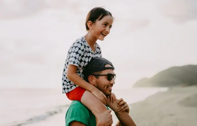 A father carrying his daughter on his shoulders at the beach.