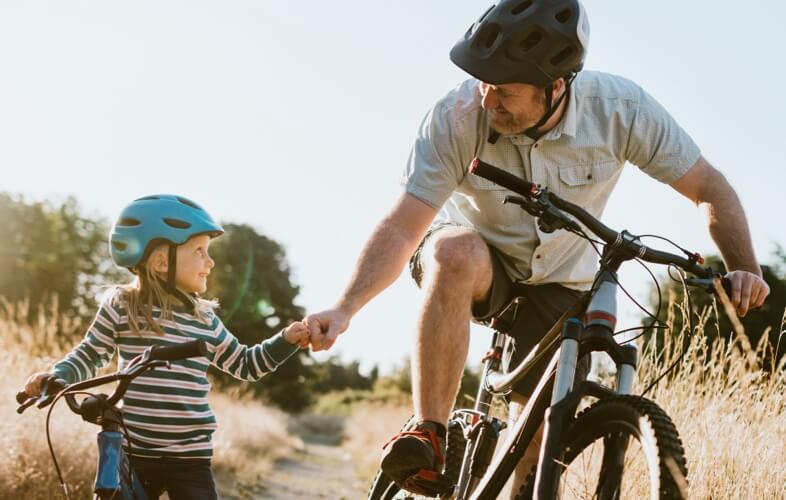 A father and young daughter riding mountain bikes on a trail together